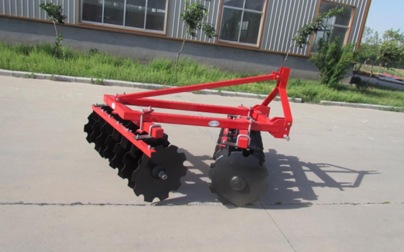 A red disc harrow with multiple discs is parked on a paved surface