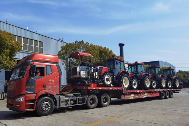 Several red tractors transported on a flatbed truck outside an industrial building