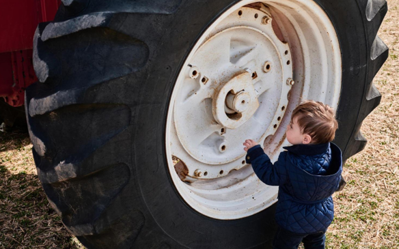 Little boy in blue beside a large tractor tire, looking curious